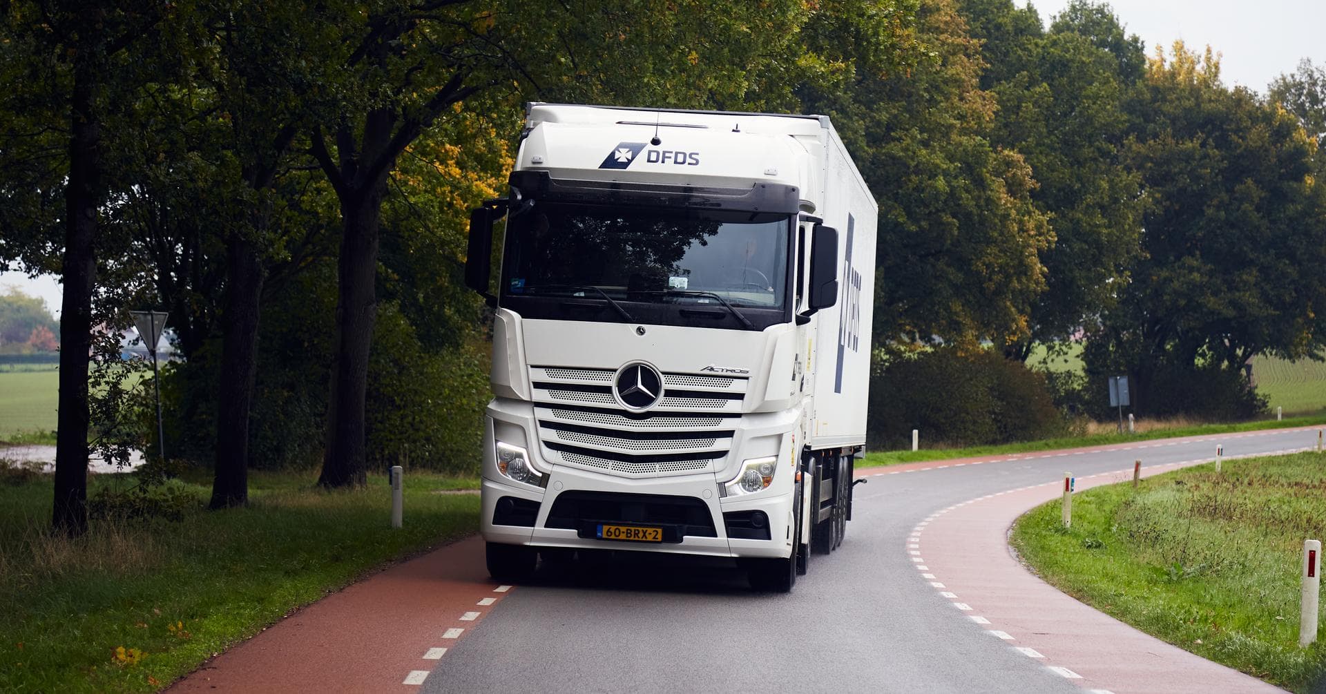 A white Dutch DFDS truck, with a white cold chain trailer behind it, is driving on a road in a rural environment