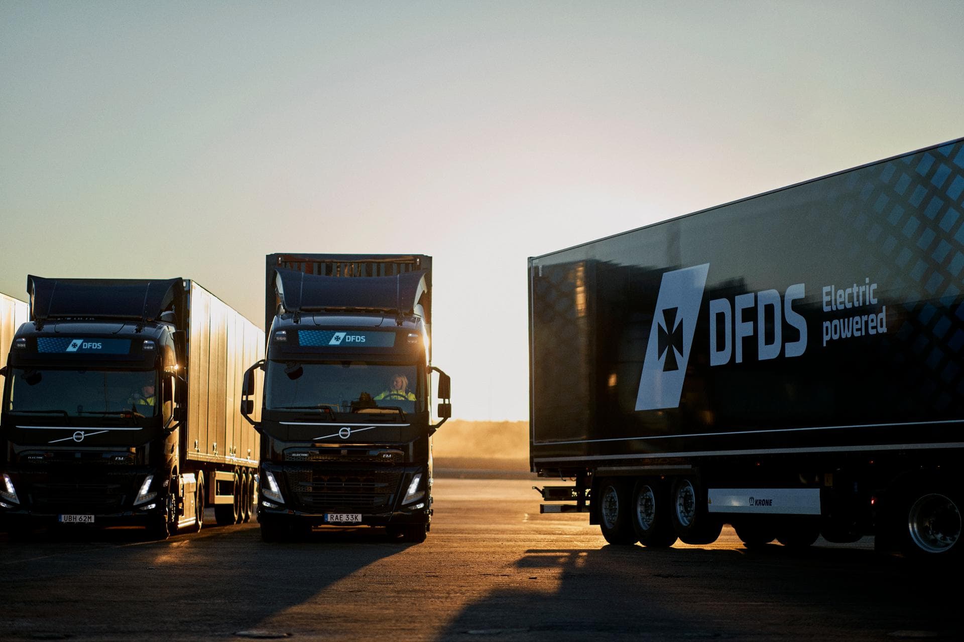 Three electric trucks in dark blue DFDS livery, standing in the sunset, standing side to side next to each other, whereas one of them is driving out of the view