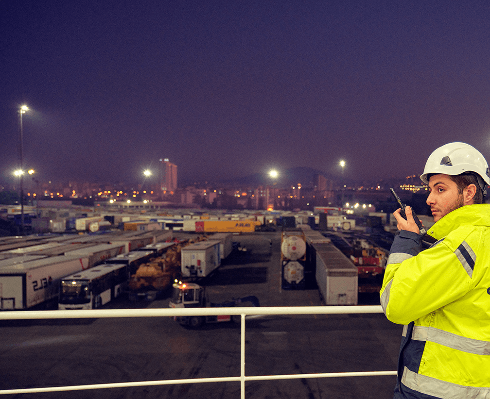 A DFDS employee wearing a high-visibility jacket and a safety helmet speaks on the intercom while overlooking the terminal at night