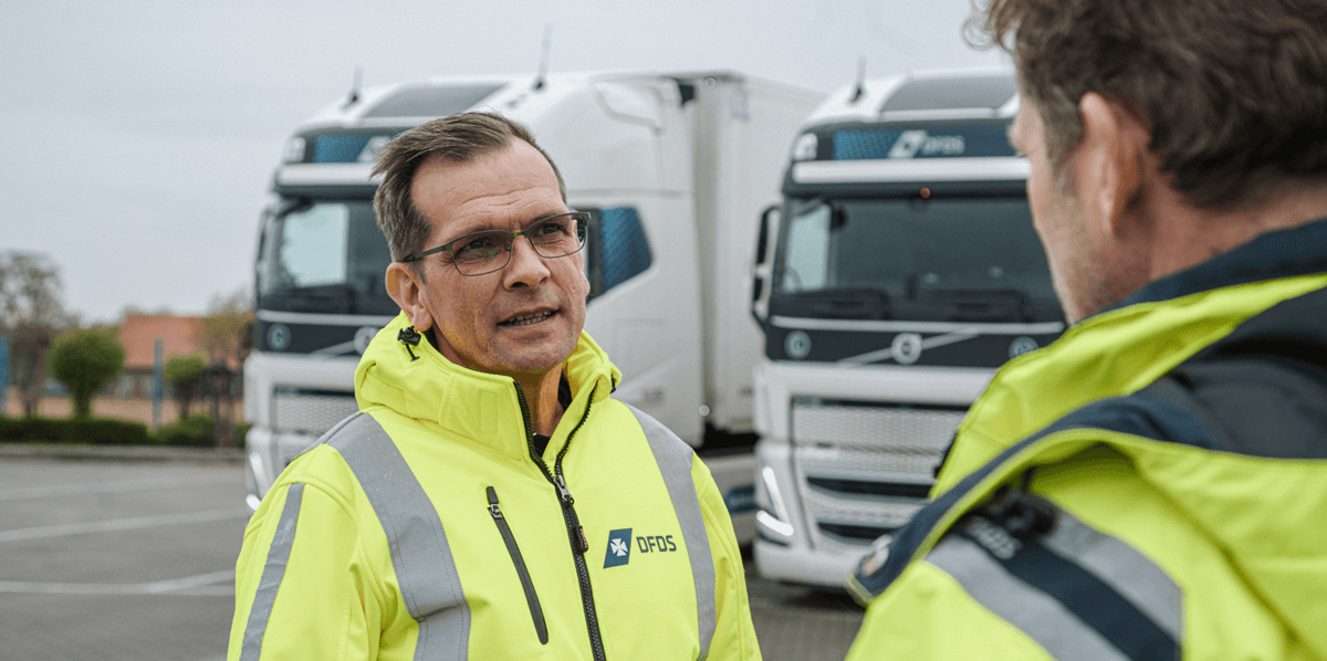 Two drivers in yellow safety jackets talking with each other, with two white DFDS-branded trucks in the background