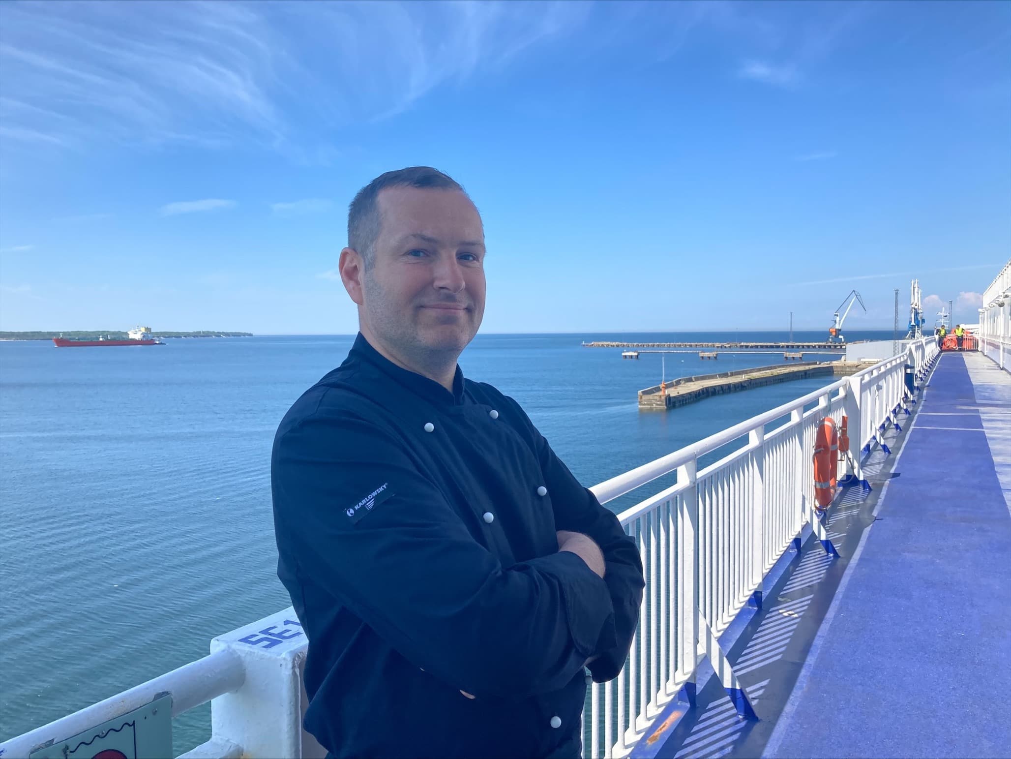 An upper body photo of the DFDS chef Argo Saar, standing on the deck of the ship PATRIA SEAWAYS, with blue skies and sea behind him