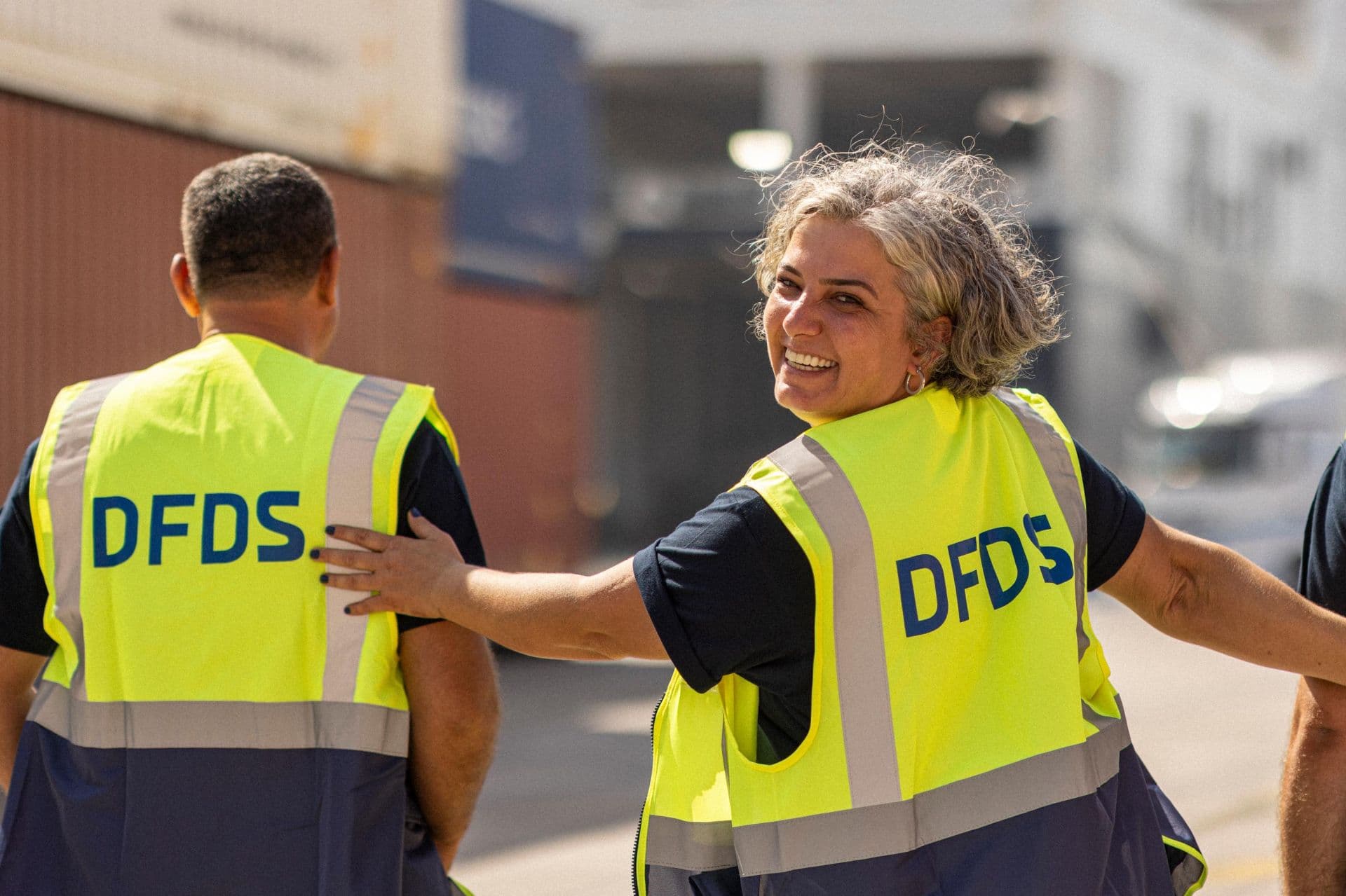 A female DFDS employee, walking between two male colleagues, wearing a yellow safety vest, while looking back and smiling!