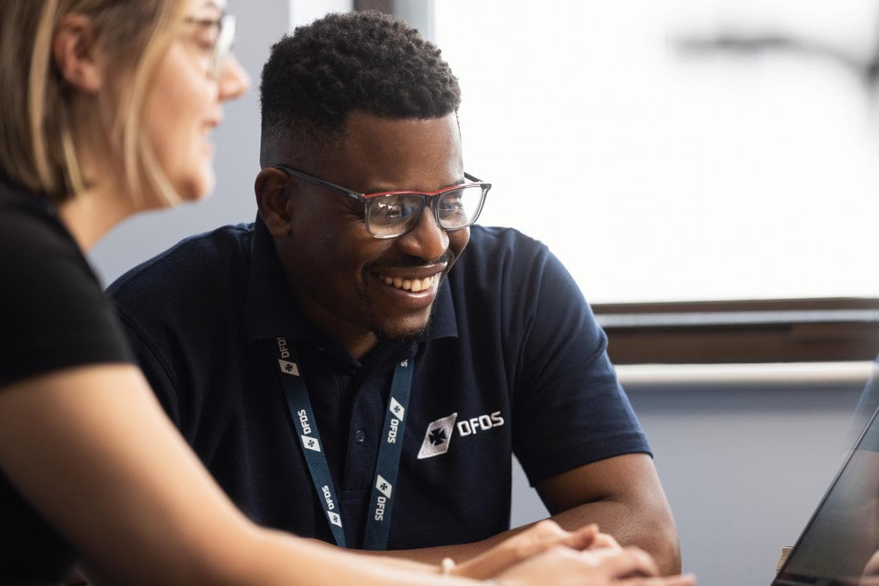 Female & male workers in dark-blue, DFDS-branded polos, sitting together behind a laptop screen, laughing
