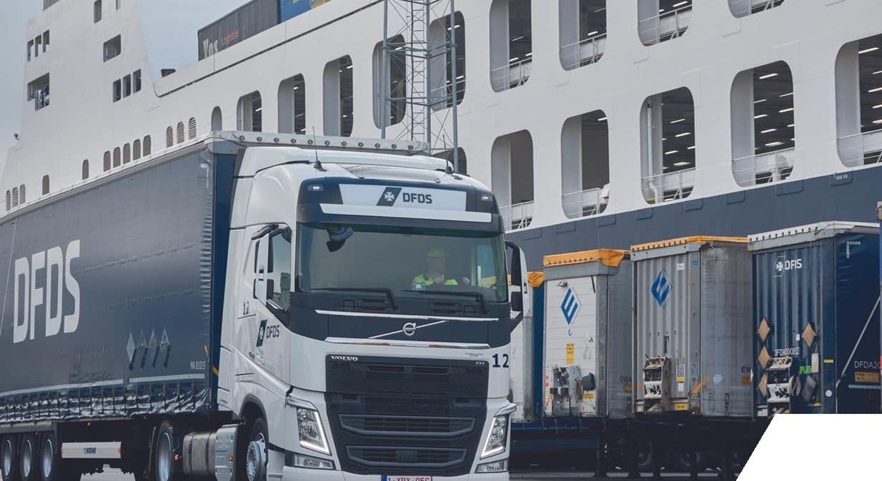 White DFDS truck with dark blue trailer, in front of DFDS vessel at bay in the harbour