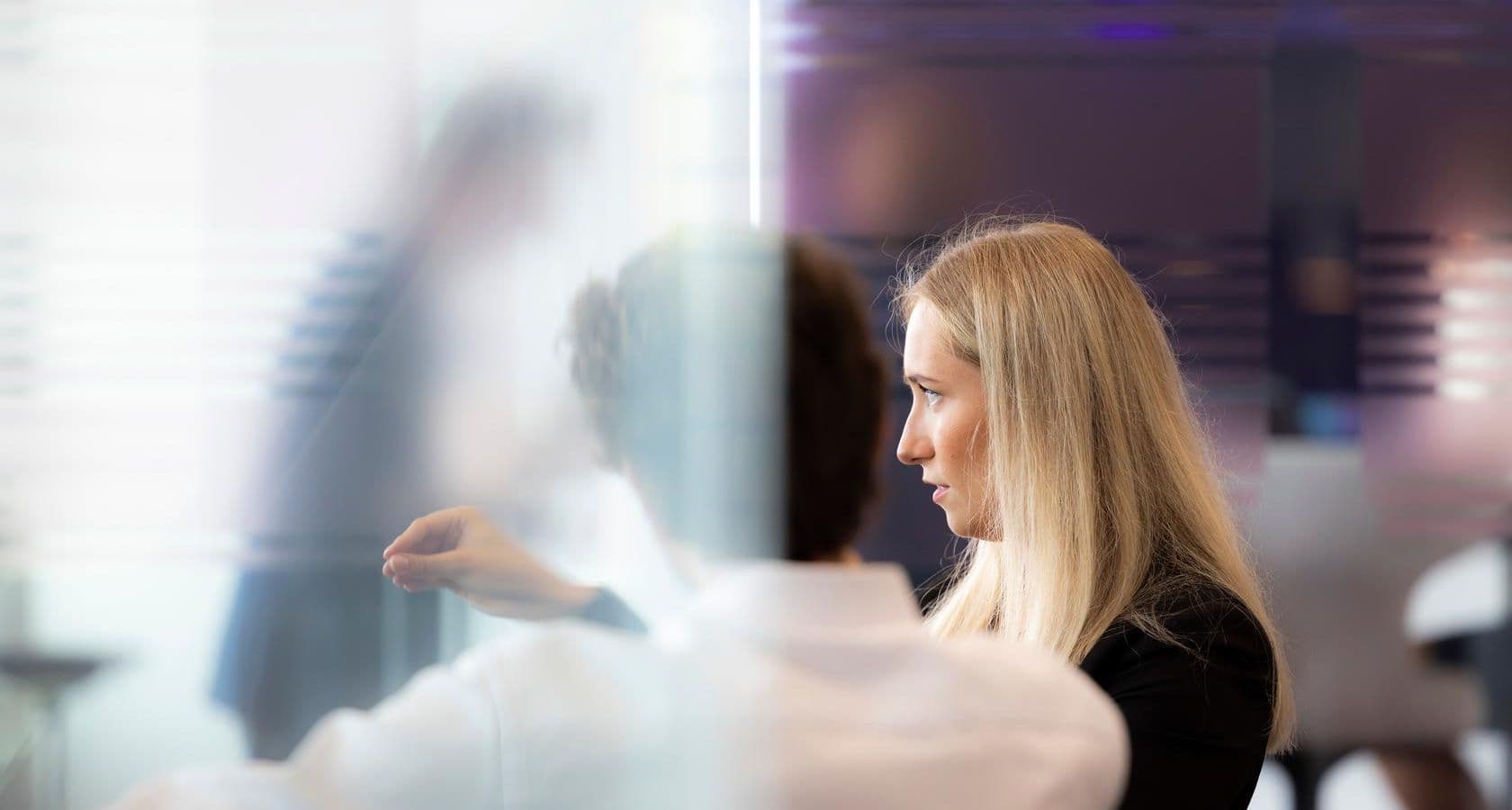 Man and woman in a meeting with other people, the man in a white blouse, seen on the back, the woman in dark clothes, pointing something out to other people in the meeting