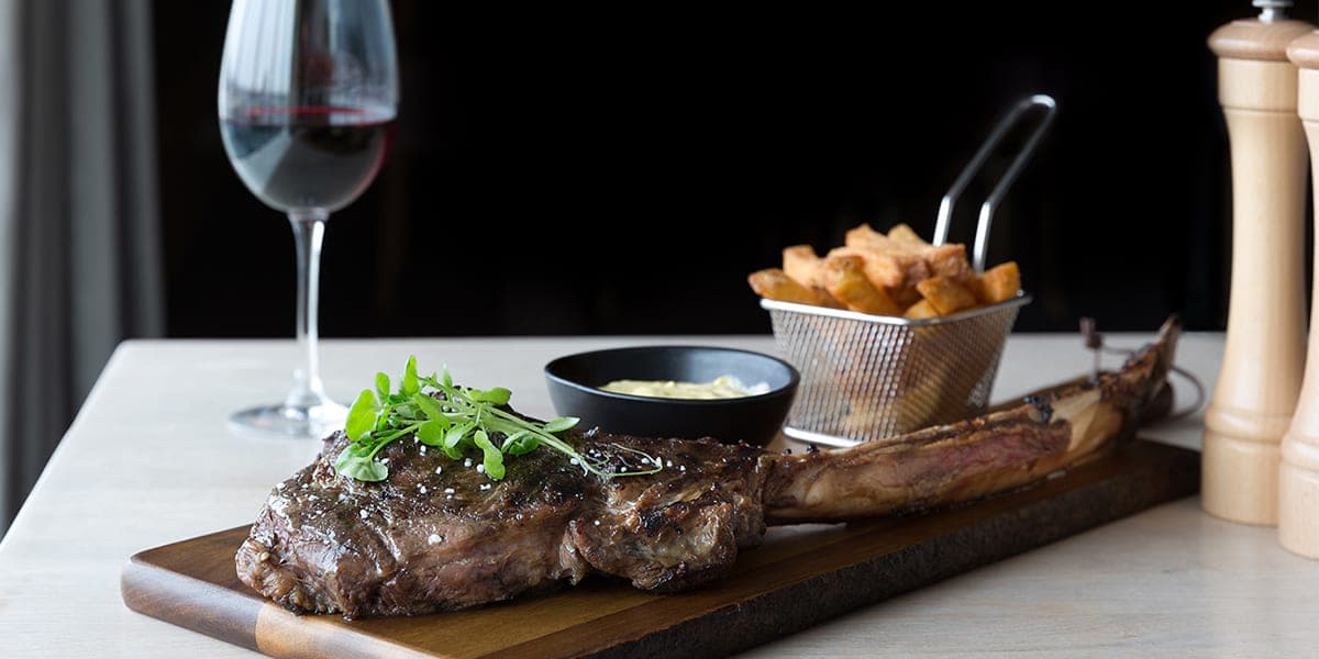 A portion of steak and chips on a wooden board with a glass of red wine on the side