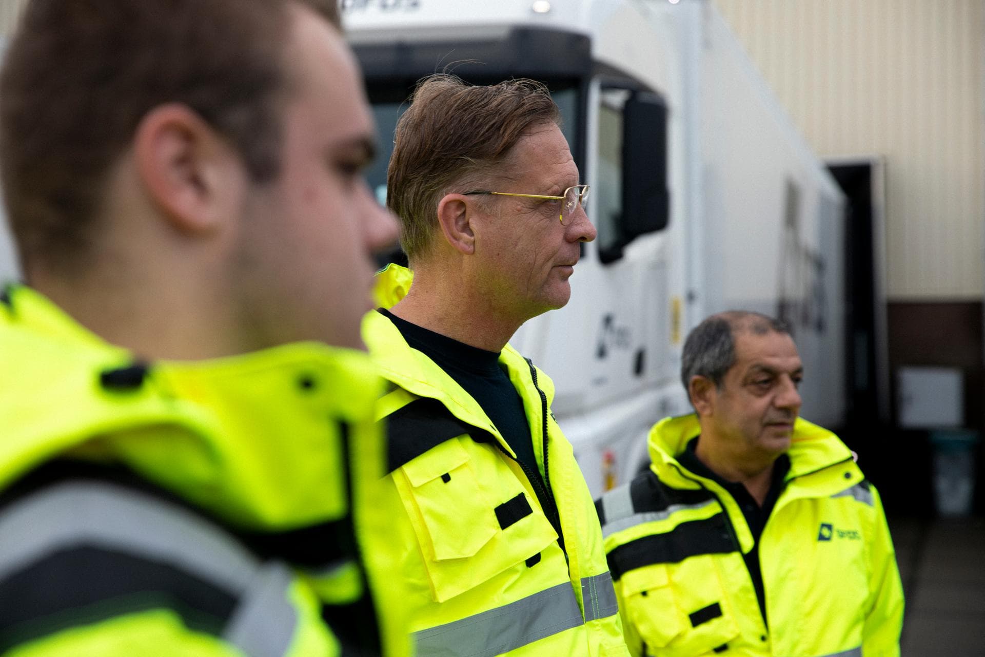 Three DFDS drivers, wearing yellow safety jackets, listening and looking and somewhat outside of the picture frame, with in the background a white cold chain truck and refrigerated trailer