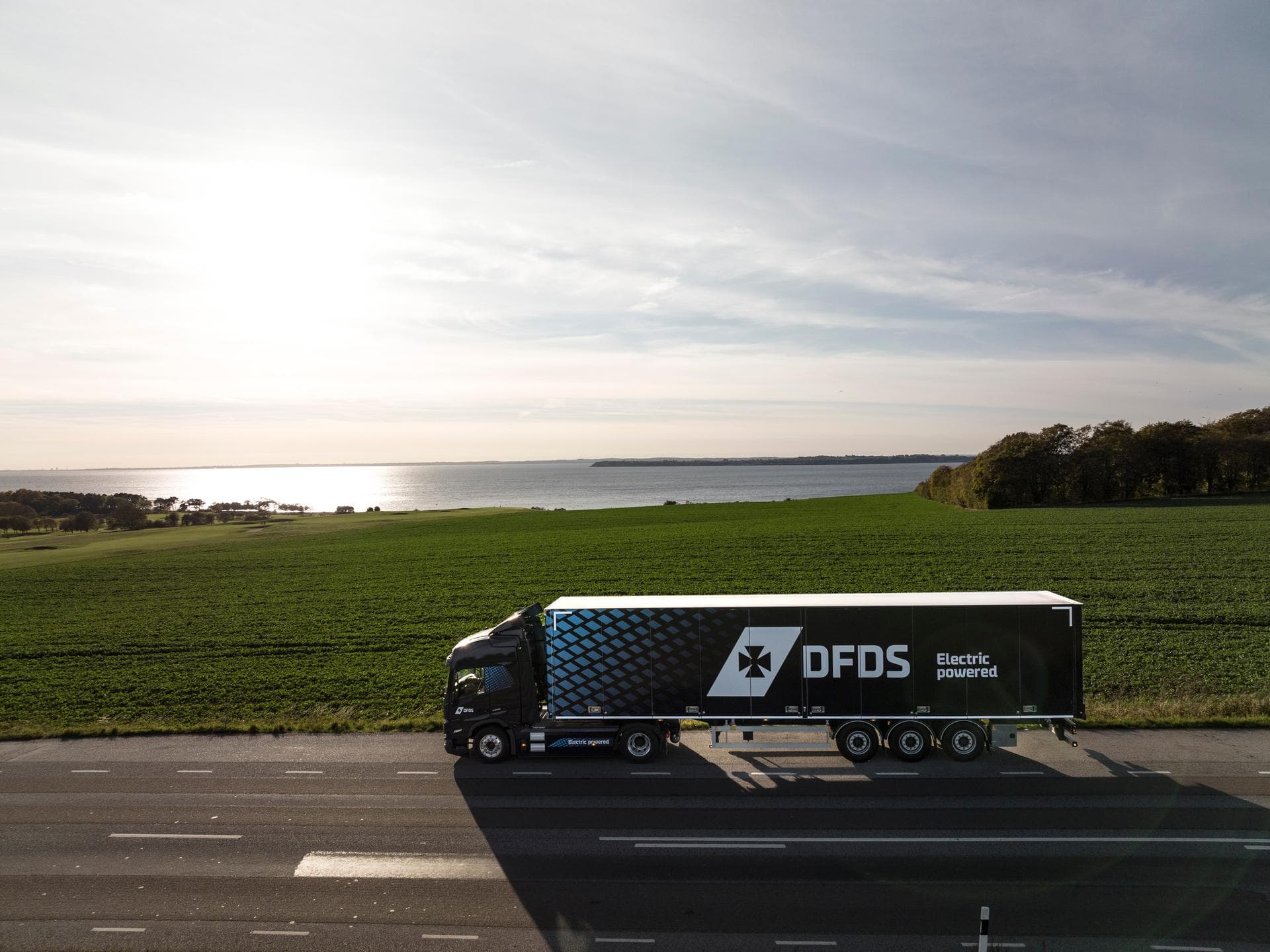 Electric truck in dark blue DFDS livery, seen from above, driving on a road in a rural & water-rich environment