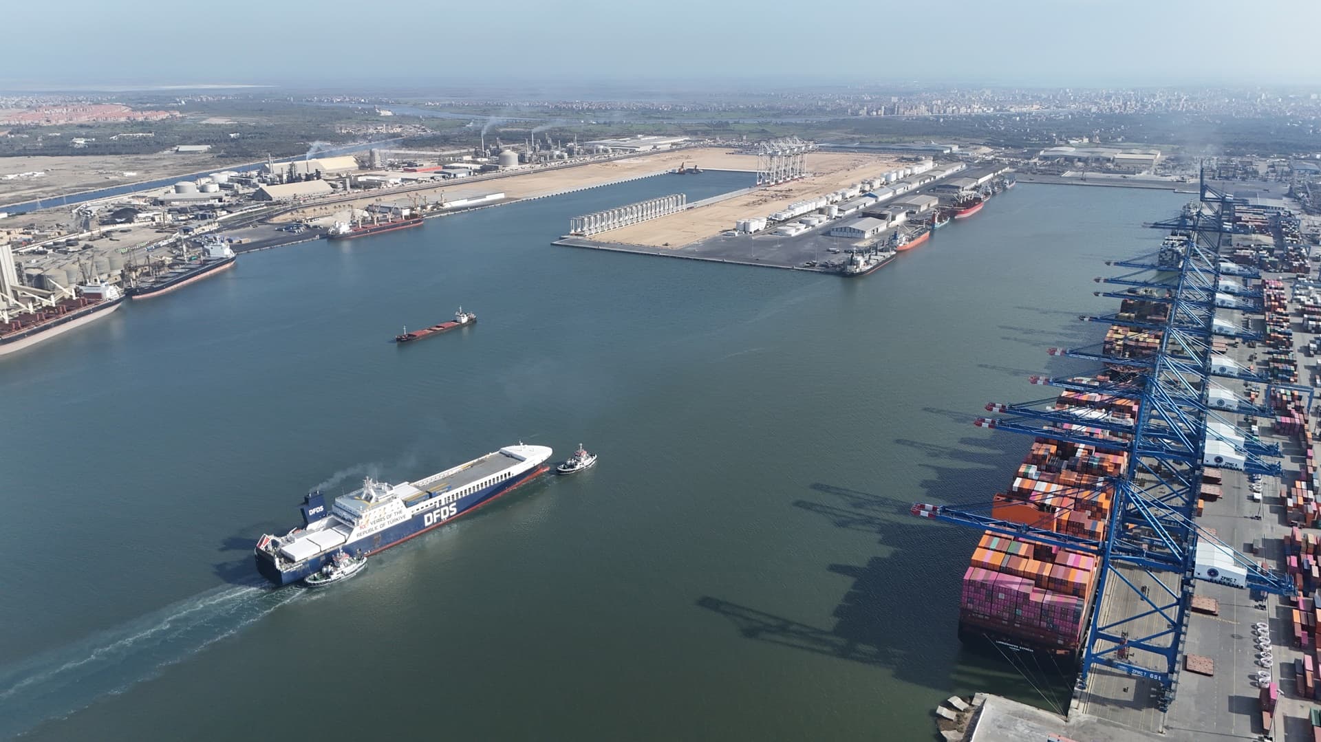 A DFDS vessel entering a harbour, accompanied by two tug boats. On the right, a long quay with container ships and cranes.