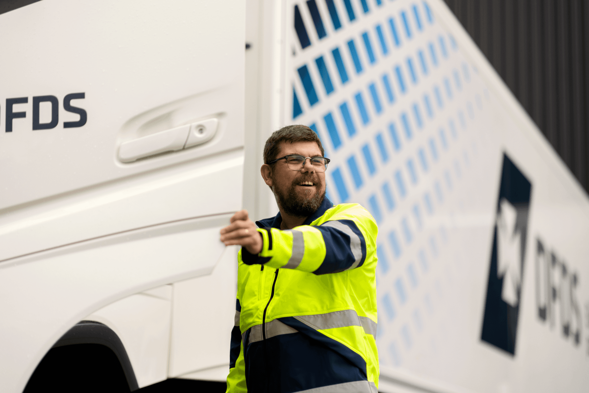 A smiling driver, wearing a safety jacket and glasses, opens the door of a white DFDS truck