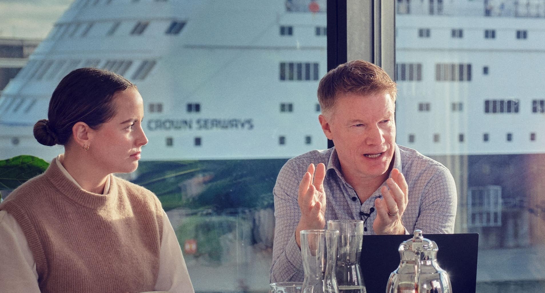 Two DFDS employees sit in a meeting room with the DFDS ferry Crown Seaways on the background