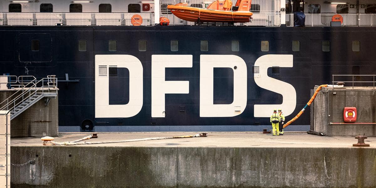 DFDS vessel lying at the quay in the harbour, seen from the side, with the white DFDS logo in the middle and two terminal workers with yellow safety jackets and wearing white helmets, standing on that quay