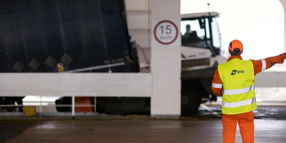 A DFDS worker with a branded, yellow safety jacket, pointing and directing a terminal truck with a trailer on the deck of a vessel