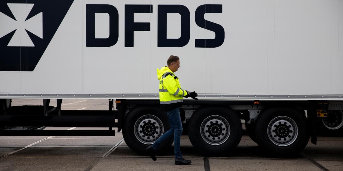 A male DFDS worker, with a yellow safety jacket, safety gloves and shoes, walking alongside a white DFDS-branded trailer